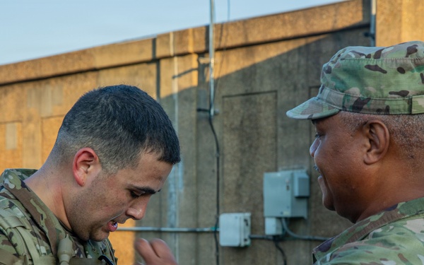 Two U.S. Army soldiers in Operational Camouflage Pattern (OCP) uniforms are engaged in what appears to be an equipment inspection or pinning ceremony outdoors near a concrete structure. The soldier on the left wears a '1st Infantry Division' shoulder patch and a rucksack, while the soldier on the right, whose name tape reads 'NUNEZ', is gesturing toward the other soldier's chest. The interaction suggests a formal or instructional moment between the two service members.