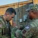 Two U.S. Army soldiers in Operational Camouflage Pattern (OCP) uniforms are engaged in what appears to be an equipment inspection or pinning ceremony outdoors near a concrete structure. The soldier on the left wears a '1st Infantry Division' shoulder patch and a rucksack, while the soldier on the right, whose name tape reads 'NUNEZ', is gesturing toward the other soldier's chest. The interaction suggests a formal or instructional moment between the two service members.