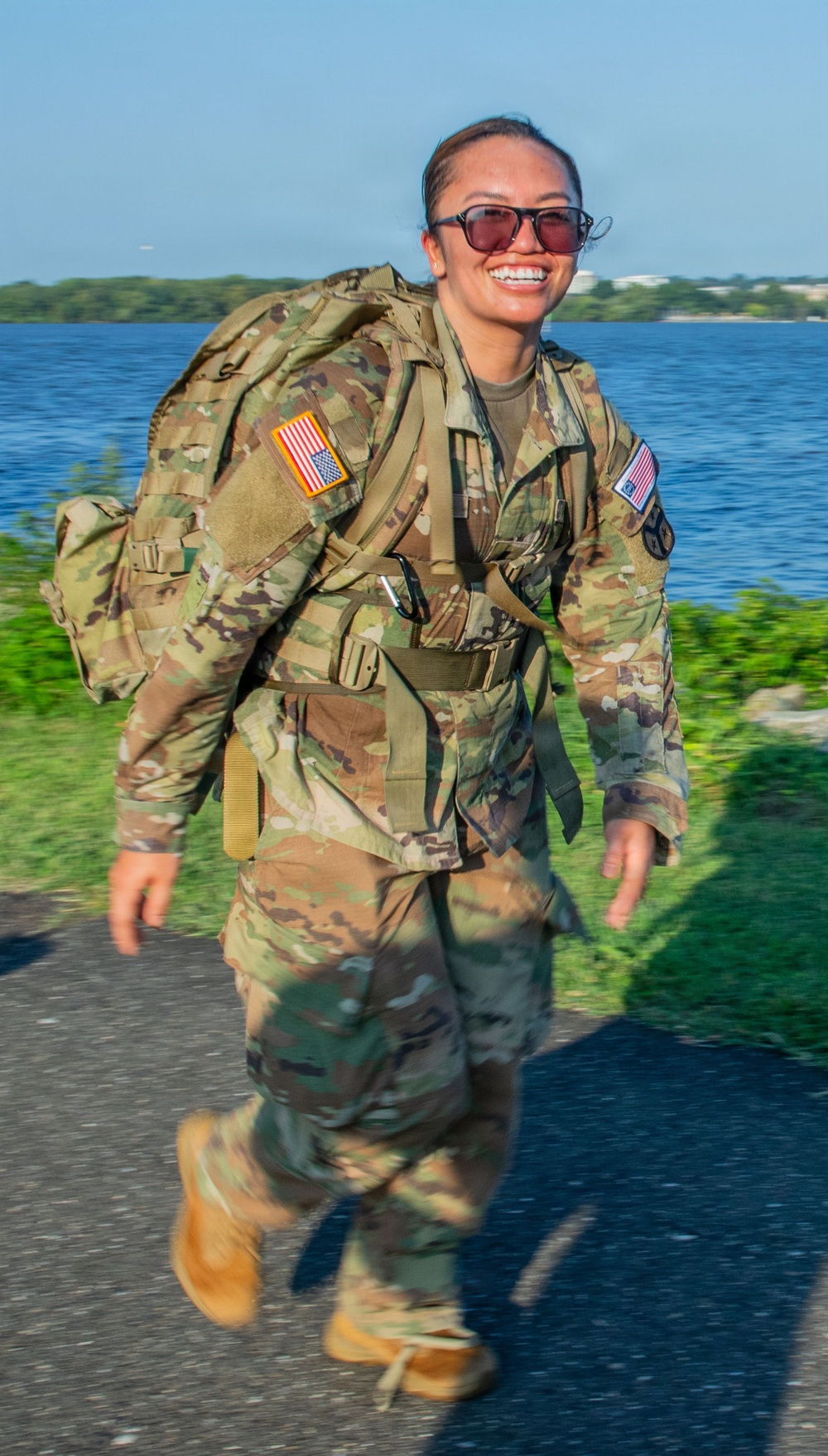 A female soldier in U.S. Army Operational Camouflage Pattern (OCP) uniform is conducting a ruck march along a paved path near a body of water. She is smiling broadly while carrying a large rucksack and wearing tinted sunglasses. The setting appears to be a waterfront area with trees and blue sky visible in the background.