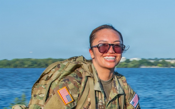 A female soldier in U.S. Army Operational Camouflage Pattern (OCP) uniform is conducting a ruck march along a paved path near a body of water. She is smiling broadly while carrying a large rucksack and wearing tinted sunglasses. The setting appears to be a waterfront area with trees and blue sky visible in the background.