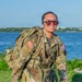A female soldier in U.S. Army Operational Camouflage Pattern (OCP) uniform is conducting a ruck march along a paved path near a body of water. She is smiling broadly while carrying a large rucksack and wearing tinted sunglasses. The setting appears to be a waterfront area with trees and blue sky visible in the background.