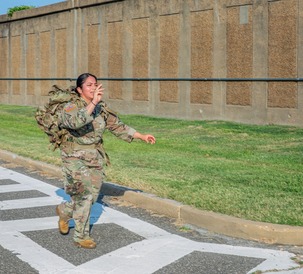 A female U.S. Army soldier in Operational Camouflage Pattern (OCP) uniform is running across a marked crosswalk while carrying a large military rucksack. She appears to be mid-stride with an expressive facial expression, possibly calling out or reacting to something. The setting appears to be a military installation based on the infrastructure visible in the background.