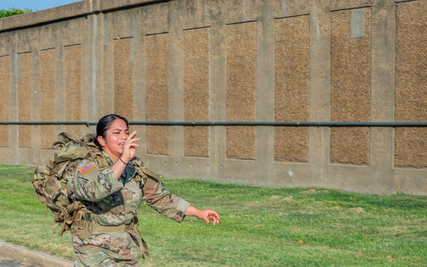 A female U.S. Army soldier in Operational Camouflage Pattern (OCP) uniform is running across a marked crosswalk while carrying a large military rucksack. She appears to be mid-stride with an expressive facial expression, possibly calling out or reacting to something. The setting appears to be a military installation based on the infrastructure visible in the background.