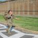 A female U.S. Army soldier in Operational Camouflage Pattern (OCP) uniform is running across a marked crosswalk while carrying a large military rucksack. She appears to be mid-stride with an expressive facial expression, possibly calling out or reacting to something. The setting appears to be a military installation based on the infrastructure visible in the background.