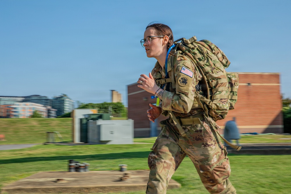 A female U.S. Army soldier in full OCP (Operational Camouflage Pattern) uniform is running or rucking at speed while carrying a large military backpack with a hydration system. She is wearing glasses and has a U.S. flag patch visible on her right shoulder, along with additional unit or qualification patches. The background shows a grassy outdoor area with brick and modern buildings, suggesting a military installation or training facility.