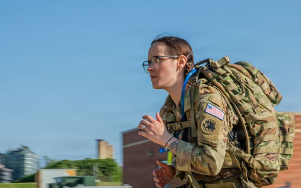 A female U.S. Army soldier in full OCP (Operational Camouflage Pattern) uniform is running or rucking at speed while carrying a large military backpack with a hydration system. She is wearing glasses and has a U.S. flag patch visible on her right shoulder, along with additional unit or qualification patches. The background shows a grassy outdoor area with brick and modern buildings, suggesting a military installation or training facility.