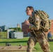 A female U.S. Army soldier in full OCP (Operational Camouflage Pattern) uniform is running or rucking at speed while carrying a large military backpack with a hydration system. She is wearing glasses and has a U.S. flag patch visible on her right shoulder, along with additional unit or qualification patches. The background shows a grassy outdoor area with brick and modern buildings, suggesting a military installation or training facility.