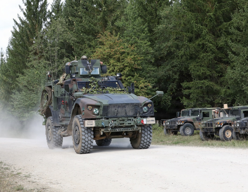 A military convoy is moving along a gravel road through a forested area. The lead vehicle is a heavily equipped Joint Light Tactical Vehicle (JLTV) with camouflage netting and a rooftop weapons system, followed by at least two Humvees parked on the roadside. The scene appears to be a military exercise or training operation based on the vehicle markings and equipment configuration.