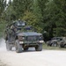 A military convoy is moving along a gravel road through a forested area. The lead vehicle is a heavily equipped Joint Light Tactical Vehicle (JLTV) with camouflage netting and a rooftop weapons system, followed by at least two Humvees parked on the roadside. The scene appears to be a military exercise or training operation based on the vehicle markings and equipment configuration.