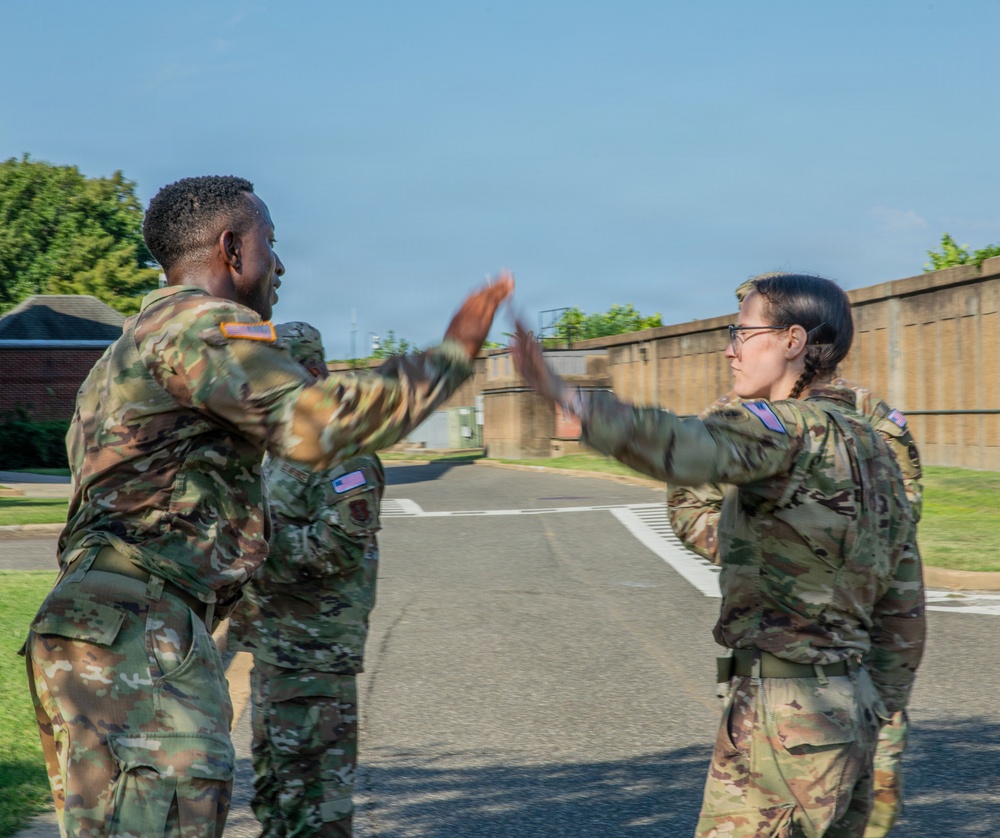 Three U.S. Army soldiers in OCP (Operational Camouflage Pattern) uniforms are engaged in what appears to be a high-five or hand gesture interaction outdoors on a military installation. The motion blur on their hands suggests active movement during the moment the photo was captured. The setting appears to be a paved area within a military base, with a concrete wall and brick building visible in the background.