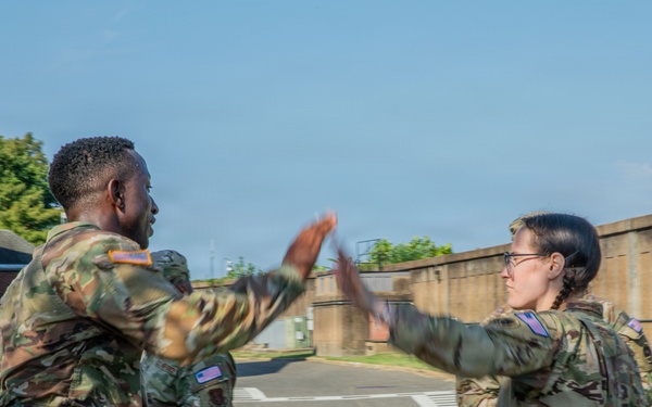 Three U.S. Army soldiers in OCP (Operational Camouflage Pattern) uniforms are engaged in what appears to be a high-five or hand gesture interaction outdoors on a military installation. The motion blur on their hands suggests active movement during the moment the photo was captured. The setting appears to be a paved area within a military base, with a concrete wall and brick building visible in the background.