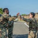 Three U.S. Army soldiers in OCP (Operational Camouflage Pattern) uniforms are engaged in what appears to be a high-five or hand gesture interaction outdoors on a military installation. The motion blur on their hands suggests active movement during the moment the photo was captured. The setting appears to be a paved area within a military base, with a concrete wall and brick building visible in the background.