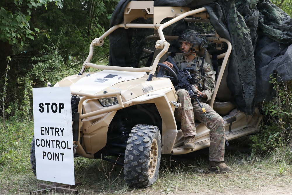 A uniformed soldier in full combat gear is seated in a tan/desert-colored military utility vehicle (UTV) at what appears to be a training or operational entry control point. A white sign reading 'STOP ENTRY CONTROL POINT' is positioned to the left of the vehicle. The setting is a wooded or semi-wooded area with camouflage netting visible behind the vehicle.