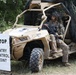 A uniformed soldier in full combat gear is seated in a tan/desert-colored military utility vehicle (UTV) at what appears to be a training or operational entry control point. A white sign reading 'STOP ENTRY CONTROL POINT' is positioned to the left of the vehicle. The setting is a wooded or semi-wooded area with camouflage netting visible behind the vehicle.
