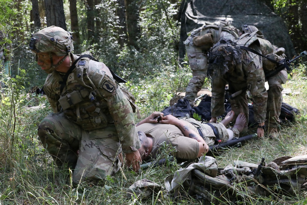 Military personnel in multicam/OCP camouflage uniforms are conducting what appears to be a combat casualty care exercise or training scenario in a wooded outdoor environment. At least two soldiers are attending to a person lying on the ground, simulating medical treatment of a casualty. The setting, equipment, and unit patches suggest this is a U.S. Army training exercise.
