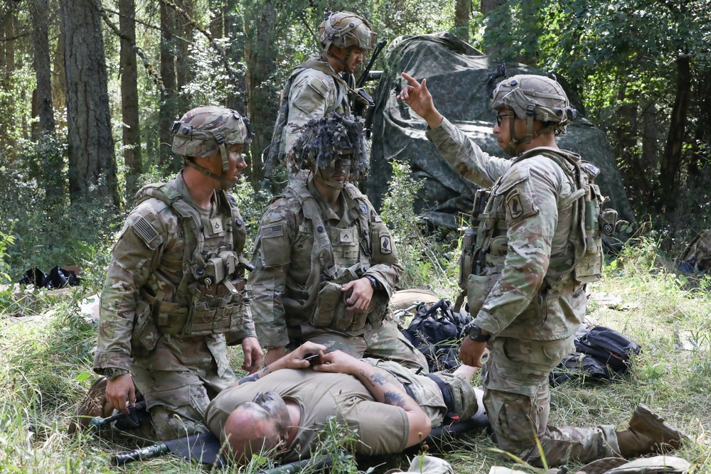 Several U.S. Army soldiers in MultiCam/OCP camouflage uniforms are conducting what appears to be a tactical combat casualty care (TCCC) training exercise in a forested area. One soldier is lying on the ground simulating a casualty while three to four others attend to him, with one soldier appearing to give instructions or directions. The setting includes a camouflage shelter/tarp in the background among tall trees.