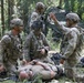 Several U.S. Army soldiers in MultiCam/OCP camouflage uniforms are conducting what appears to be a tactical combat casualty care (TCCC) training exercise in a forested area. One soldier is lying on the ground simulating a casualty while three to four others attend to him, with one soldier appearing to give instructions or directions. The setting includes a camouflage shelter/tarp in the background among tall trees.