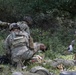 The image depicts what appears to be a military medical training exercise, with several soldiers in camouflage uniforms attending to simulated casualties on the ground. Multiple personnel are kneeling and providing care to individuals lying in a grassy, wooded area. The presence of training materials, simulated wounds, and the organized nature of the scene strongly suggests a TCCC (Tactical Combat Casualty Care) or mass casualty training drill.