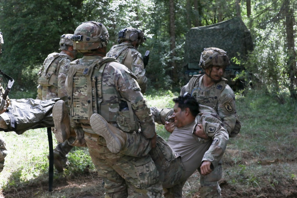 The image shows U.S. Army soldiers in Operational Camouflage Pattern (OCP) uniforms conducting a casualty evacuation (CASEVAC) drill or training exercise in a wooded outdoor environment. Multiple soldiers are carrying a simulated casualty, with one soldier bearing the person on his back while others assist. A camouflage-netted shelter or vehicle is visible in the background.