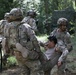 The image shows U.S. Army soldiers in Operational Camouflage Pattern (OCP) uniforms conducting a casualty evacuation (CASEVAC) drill or training exercise in a wooded outdoor environment. Multiple soldiers are carrying a simulated casualty, with one soldier bearing the person on his back while others assist. A camouflage-netted shelter or vehicle is visible in the background.