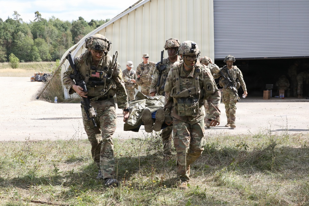 U.S. Army soldiers in full combat gear are conducting a casualty evacuation drill, with two soldiers carrying an incapacitated person on a litter or improvised stretcher. The scene appears to be a military training exercise at a base or installation, with additional soldiers visible in the background near a large metal building. Soldiers are wearing OCP (Operational Camouflage Pattern) uniforms with American flag patches and full tactical equipment.