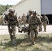 U.S. Army soldiers in full combat gear are conducting a casualty evacuation drill, with two soldiers carrying an incapacitated person on a litter or improvised stretcher. The scene appears to be a military training exercise at a base or installation, with additional soldiers visible in the background near a large metal building. Soldiers are wearing OCP (Operational Camouflage Pattern) uniforms with American flag patches and full tactical equipment.
