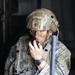 A U.S. Army soldier in full combat gear is communicating via radio handset. The soldier wears MultiCam (OCP) camouflage uniform with visible unit patches including Ranger and Airborne tabs, and has face camouflage paint applied. The image appears to be taken during a field training exercise or operational deployment.