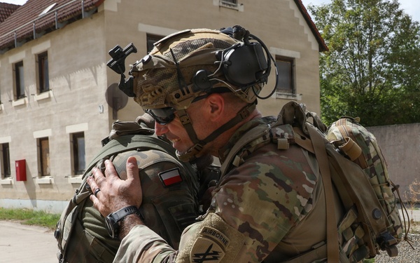Two soldiers in full combat gear are embracing in what appears to be a friendly greeting or farewell. One soldier wears a Polish flag patch on their shoulder, while the other displays a US Army 'Airborne' patch and multicam uniform, suggesting an allied military interaction. The setting appears to be a military installation or training area in Europe.