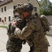 Two soldiers in full combat gear are embracing in what appears to be a friendly greeting or farewell. One soldier wears a Polish flag patch on their shoulder, while the other displays a US Army 'Airborne' patch and multicam uniform, suggesting an allied military interaction. The setting appears to be a military installation or training area in Europe.