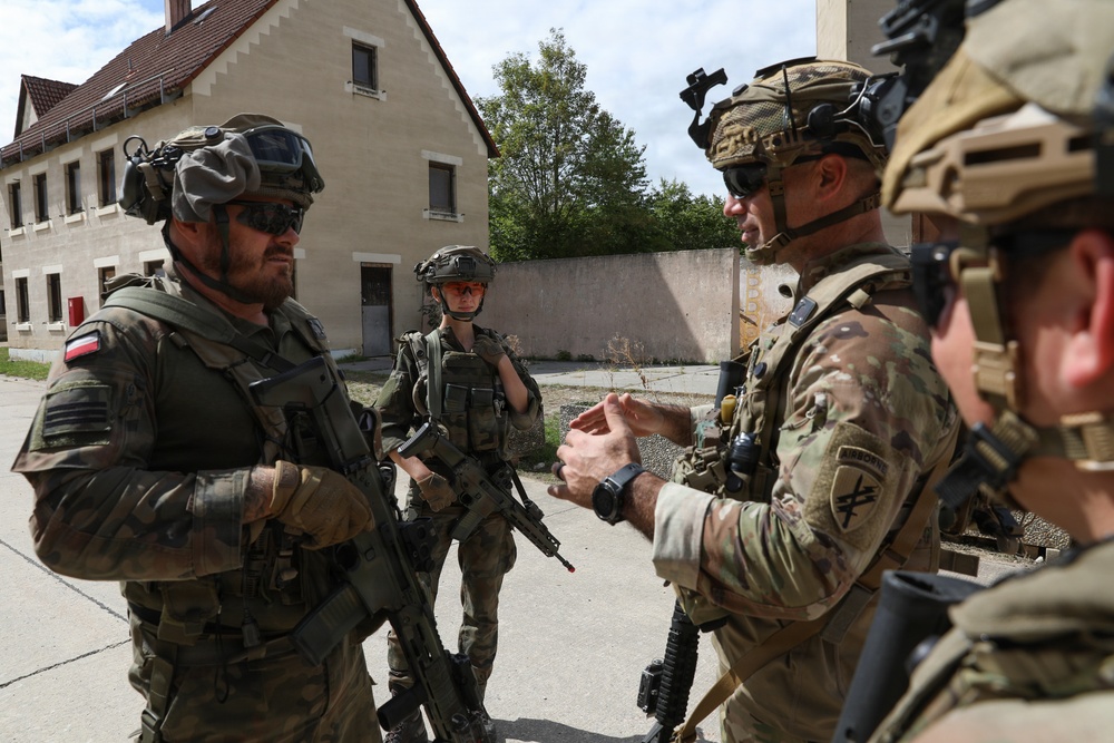 The image shows a group of military personnel in full combat gear engaged in what appears to be a briefing or coordination discussion in an outdoor urban training environment. At least two different national forces are represented, with one soldier visibly wearing a Polish flag patch on the left arm. The setting appears to be a military training facility or mock urban environment.