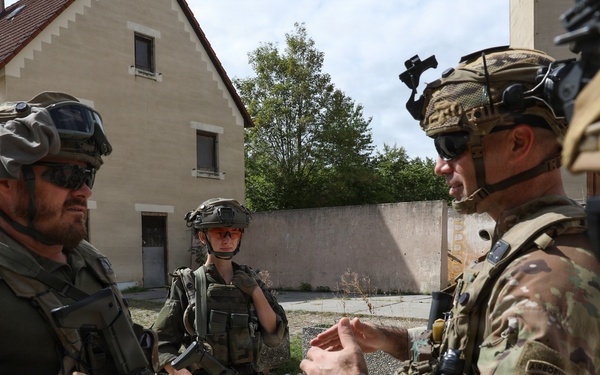 The image shows a group of military personnel in full combat gear engaged in what appears to be a briefing or coordination discussion in an outdoor urban training environment. At least two different national forces are represented, with one soldier visibly wearing a Polish flag patch on the left arm. The setting appears to be a military training facility or mock urban environment.