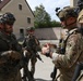 The image shows a group of military personnel in full combat gear engaged in what appears to be a briefing or coordination discussion in an outdoor urban training environment. At least two different national forces are represented, with one soldier visibly wearing a Polish flag patch on the left arm. The setting appears to be a military training facility or mock urban environment.