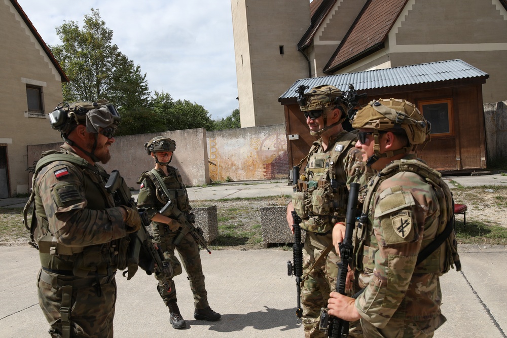 Four armed soldiers in full combat gear are gathered in an outdoor courtyard area between buildings, appearing to be in conversation. Two soldiers on the left wear Polish military patches (red and white flag visible), while two on the right wear what appears to be US Army uniforms with multicam pattern and unit patches. The setting suggests a joint military training exercise or operational briefing.