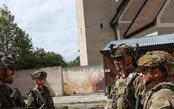 Four armed soldiers in full combat gear are gathered in an outdoor courtyard area between buildings, appearing to be in conversation. Two soldiers on the left wear Polish military patches (red and white flag visible), while two on the right wear what appears to be US Army uniforms with multicam pattern and unit patches. The setting suggests a joint military training exercise or operational briefing.