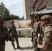 Four armed soldiers in full combat gear are gathered in an outdoor courtyard area between buildings, appearing to be in conversation. Two soldiers on the left wear Polish military patches (red and white flag visible), while two on the right wear what appears to be US Army uniforms with multicam pattern and unit patches. The setting suggests a joint military training exercise or operational briefing.