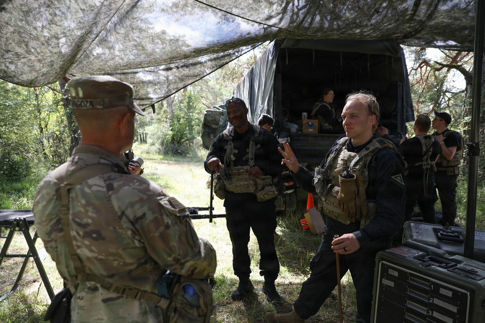 A group of military or paramilitary personnel are gathered in a wooded outdoor area beneath a camouflage netting canopy near a covered military truck. One soldier in multicam uniform with a visible name tape faces several individuals wearing dark tactical vests and gear. The scene appears to be a field briefing, staging area, or training exercise.