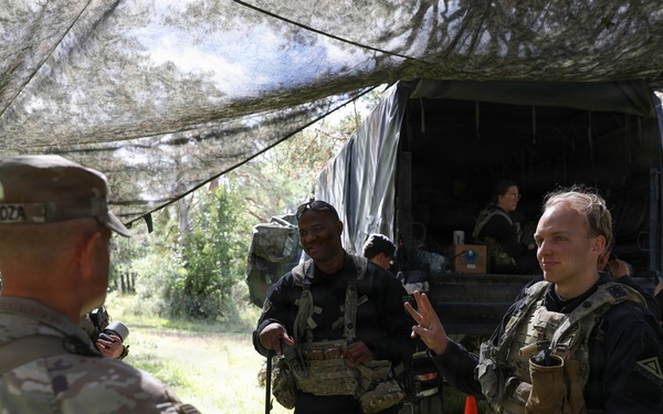 A group of military or paramilitary personnel are gathered in a wooded outdoor area beneath a camouflage netting canopy near a covered military truck. One soldier in multicam uniform with a visible name tape faces several individuals wearing dark tactical vests and gear. The scene appears to be a field briefing, staging area, or training exercise.