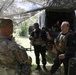 A group of military or paramilitary personnel are gathered in a wooded outdoor area beneath a camouflage netting canopy near a covered military truck. One soldier in multicam uniform with a visible name tape faces several individuals wearing dark tactical vests and gear. The scene appears to be a field briefing, staging area, or training exercise.