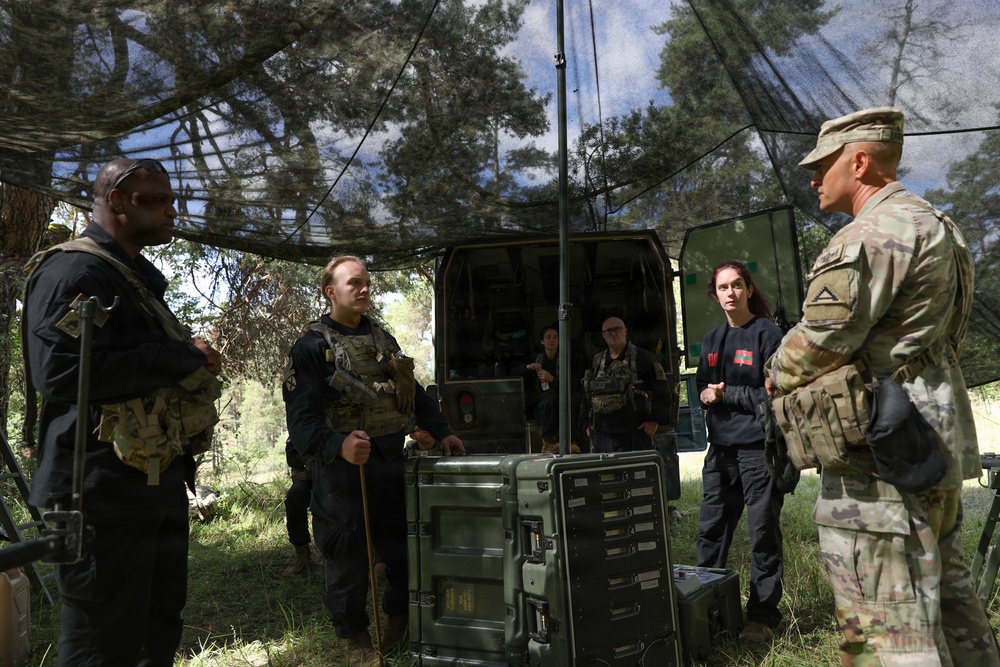 A group of individuals in tactical/military gear are gathered under a camouflage netting canopy in an outdoor wooded setting. A soldier in US Army MultiCam (OCP) uniform appears to be briefing or instructing the group near military equipment cases and a vehicle. The setting suggests a field exercise, training operation, or tactical briefing scenario.
