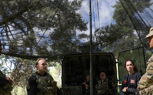 A group of individuals in tactical/military gear are gathered under a camouflage netting canopy in an outdoor wooded setting. A soldier in US Army MultiCam (OCP) uniform appears to be briefing or instructing the group near military equipment cases and a vehicle. The setting suggests a field exercise, training operation, or tactical briefing scenario.