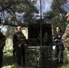 A group of individuals in tactical/military gear are gathered under a camouflage netting canopy in an outdoor wooded setting. A soldier in US Army MultiCam (OCP) uniform appears to be briefing or instructing the group near military equipment cases and a vehicle. The setting suggests a field exercise, training operation, or tactical briefing scenario.