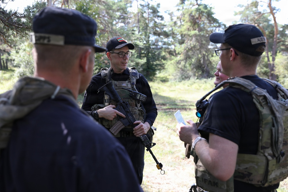A group of military or law enforcement personnel are gathered outdoors in a forested area, engaged in a briefing or discussion. The central figure is armed with a rifle and wearing a tactical vest with a flag patch visible on his cap. At least three individuals are present, with cap labels suggesting multinational participation.