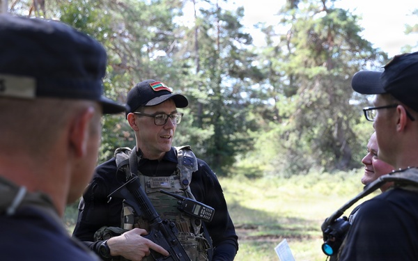 A group of military or law enforcement personnel are gathered outdoors in a forested area, engaged in a briefing or discussion. The central figure is armed with a rifle and wearing a tactical vest with a flag patch visible on his cap. At least three individuals are present, with cap labels suggesting multinational participation.