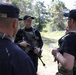 A group of military or law enforcement personnel are gathered outdoors in a forested area, engaged in a briefing or discussion. The central figure is armed with a rifle and wearing a tactical vest with a flag patch visible on his cap. At least three individuals are present, with cap labels suggesting multinational participation.