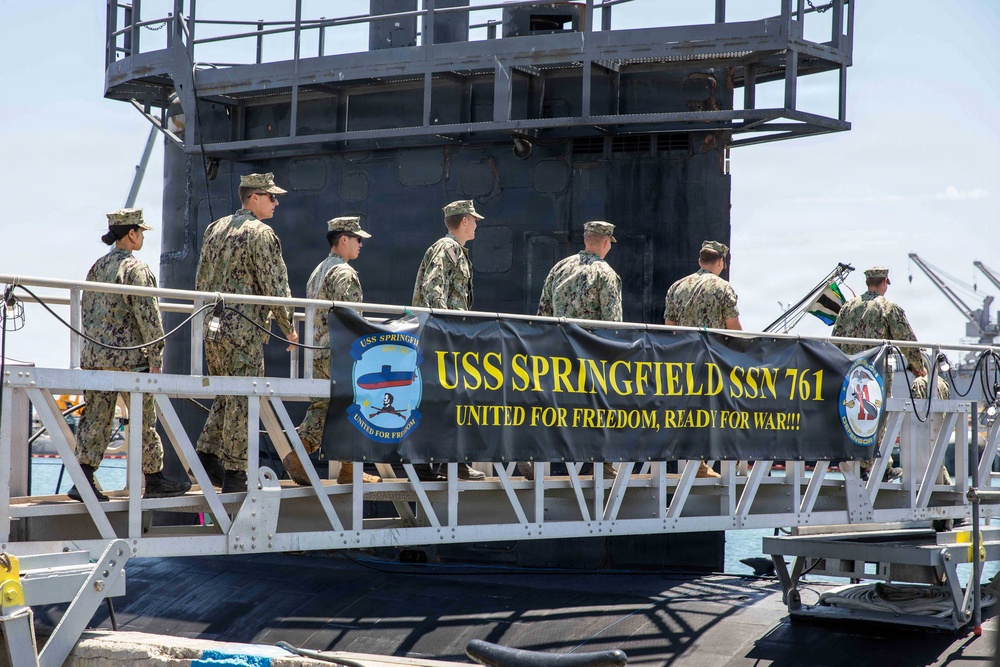 The image shows U.S. Navy personnel in camouflage uniforms boarding or standing on the USS Springfield SSN 761, a Los Angeles-class attack submarine. A prominent banner on the gangway reads 'USS SPRINGFIELD SSN 761 - UNITED FOR FREEDOM, READY FOR WAR!!!' along with the ship's insignia. The scene appears to be at a naval port or pier during daylight hours.