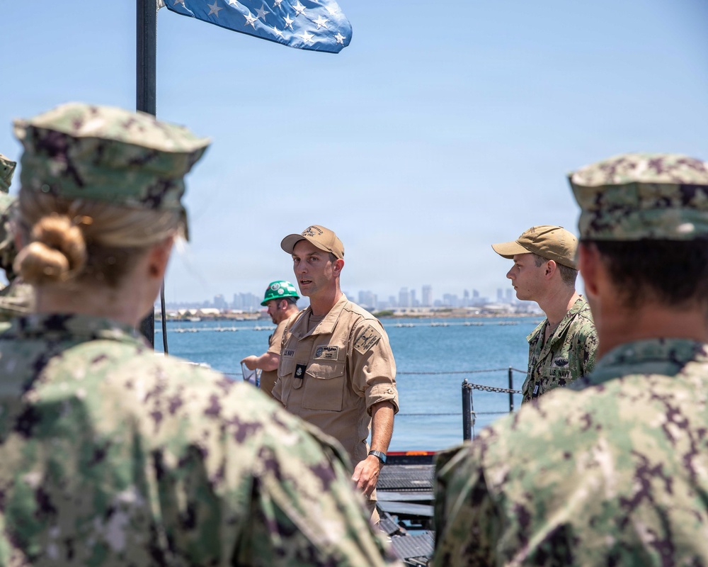 A group of U.S. Navy personnel are gathered outdoors near a waterfront, with one individual in a tan uniform and cap appearing to address or brief the group. The setting appears to be a naval installation or pier, with a body of water and a city skyline visible in the background. An American flag is partially visible at the top of the frame.