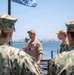 A group of U.S. Navy personnel are gathered outdoors near a waterfront, with one individual in a tan uniform and cap appearing to address or brief the group. The setting appears to be a naval installation or pier, with a body of water and a city skyline visible in the background. An American flag is partially visible at the top of the frame.