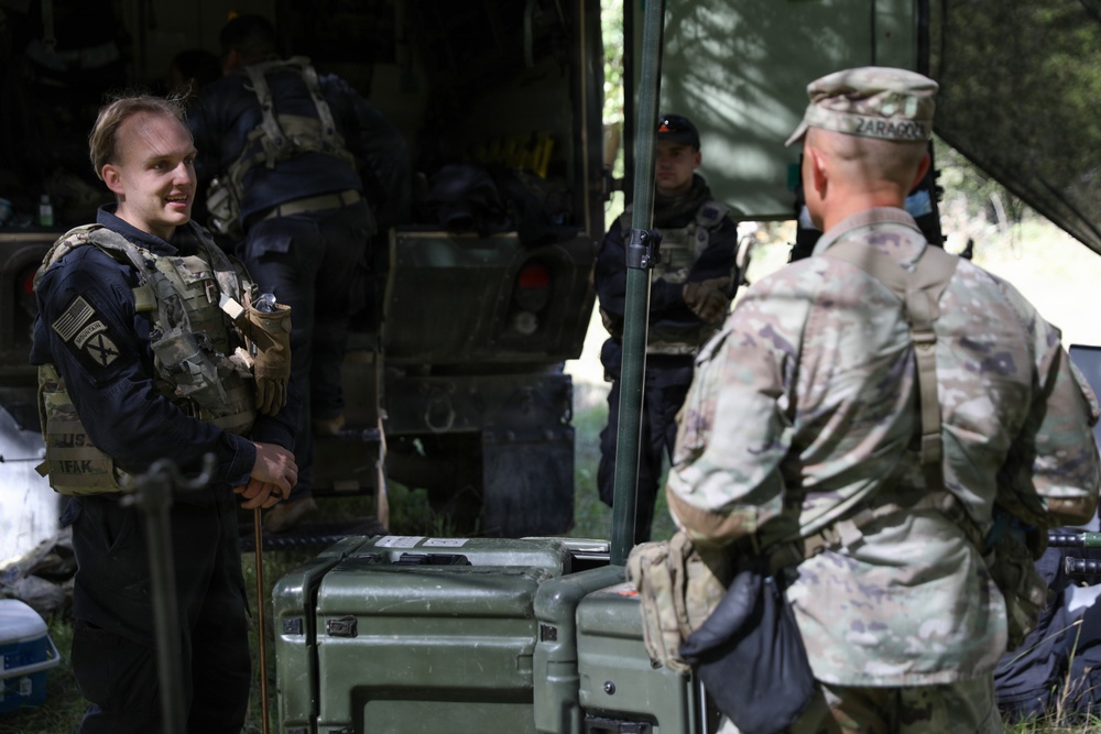 The image shows military personnel in a field setting, appearing to be engaged in a briefing or conversation near a military vehicle and equipment. At least three individuals are visible, wearing tactical gear and uniforms, with one person in MultiCam/OCP camouflage pattern and another in dark clothing with a US flag patch. The setting appears to be an outdoor staging or operations area with military cases and equipment visible.