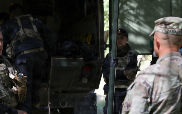 The image shows military personnel in a field setting, appearing to be engaged in a briefing or conversation near a military vehicle and equipment. At least three individuals are visible, wearing tactical gear and uniforms, with one person in MultiCam/OCP camouflage pattern and another in dark clothing with a US flag patch. The setting appears to be an outdoor staging or operations area with military cases and equipment visible.
