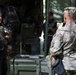 The image shows military personnel in a field setting, appearing to be engaged in a briefing or conversation near a military vehicle and equipment. At least three individuals are visible, wearing tactical gear and uniforms, with one person in MultiCam/OCP camouflage pattern and another in dark clothing with a US flag patch. The setting appears to be an outdoor staging or operations area with military cases and equipment visible.